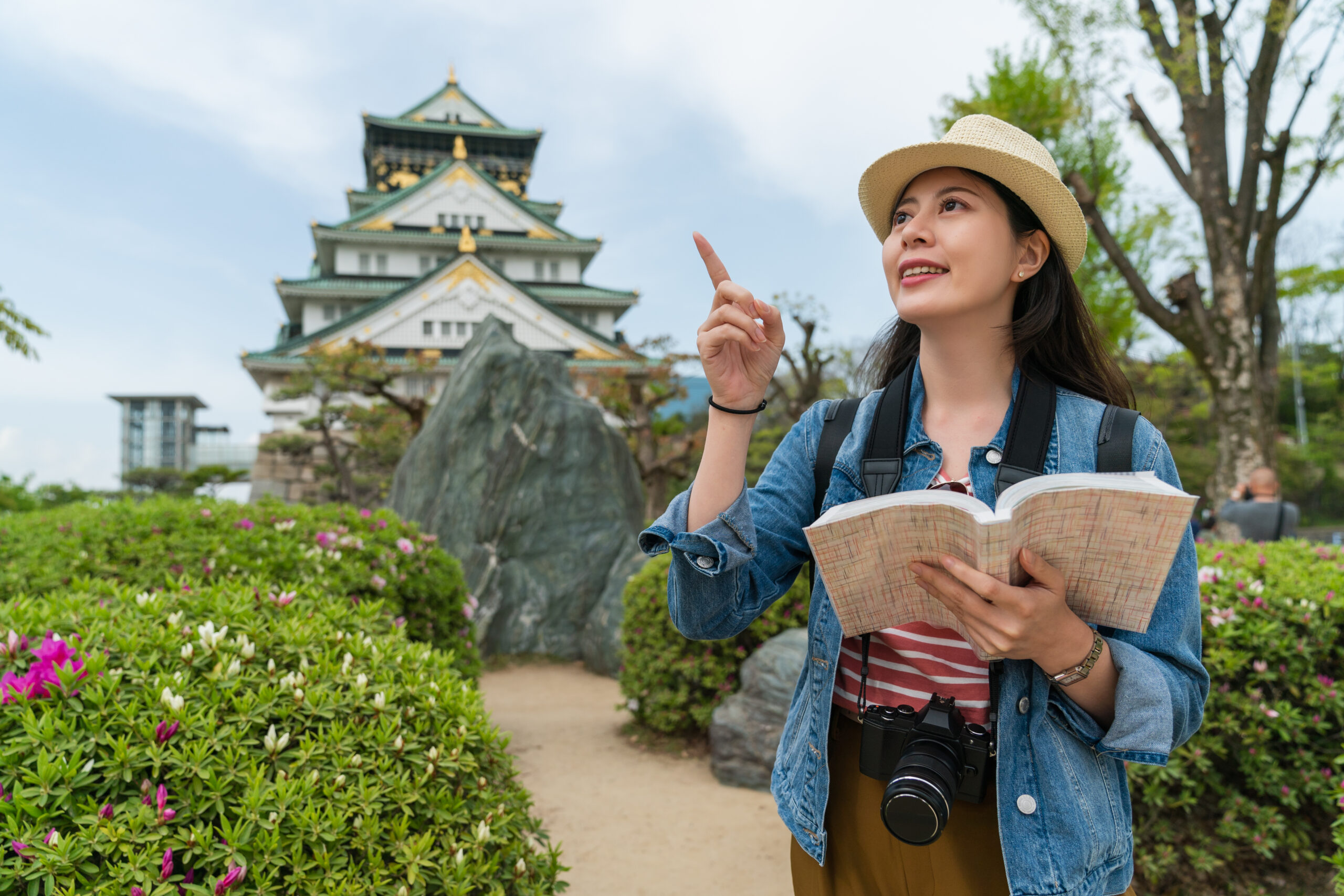 smiling Taiwanese girl backpacker finger pointing into space at a landmark while visiting Osaka castle park using travel guide at springtime in japan